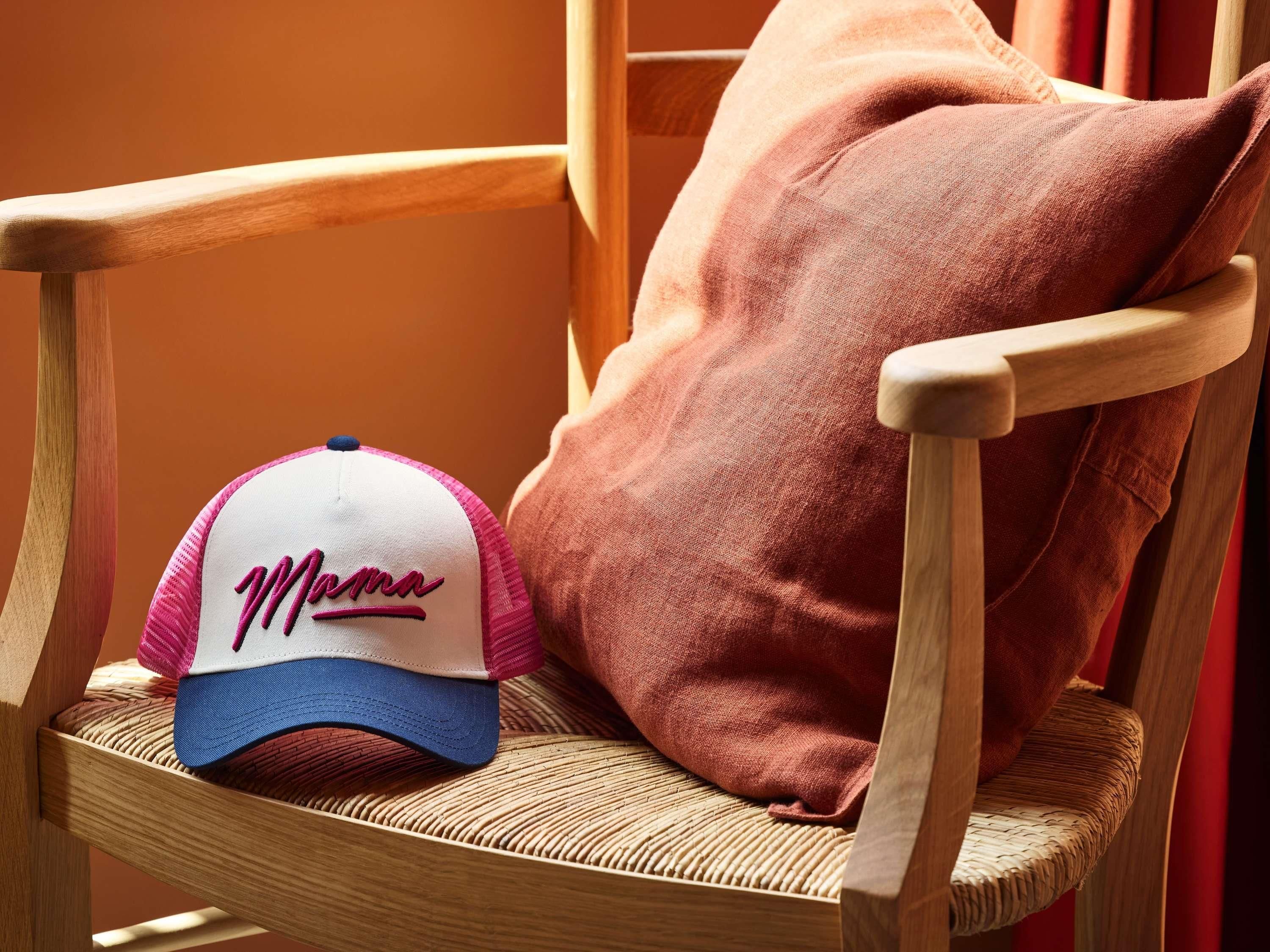 Una silla de madera con asiento y respaldo tejido, un cojín rosa y una gorra de béisbol con el bordado 'Mamma'.