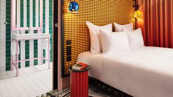 A hotel room with a bed, a bedside table with a book, a mirror, a red trash bin, and a patterned carpet.