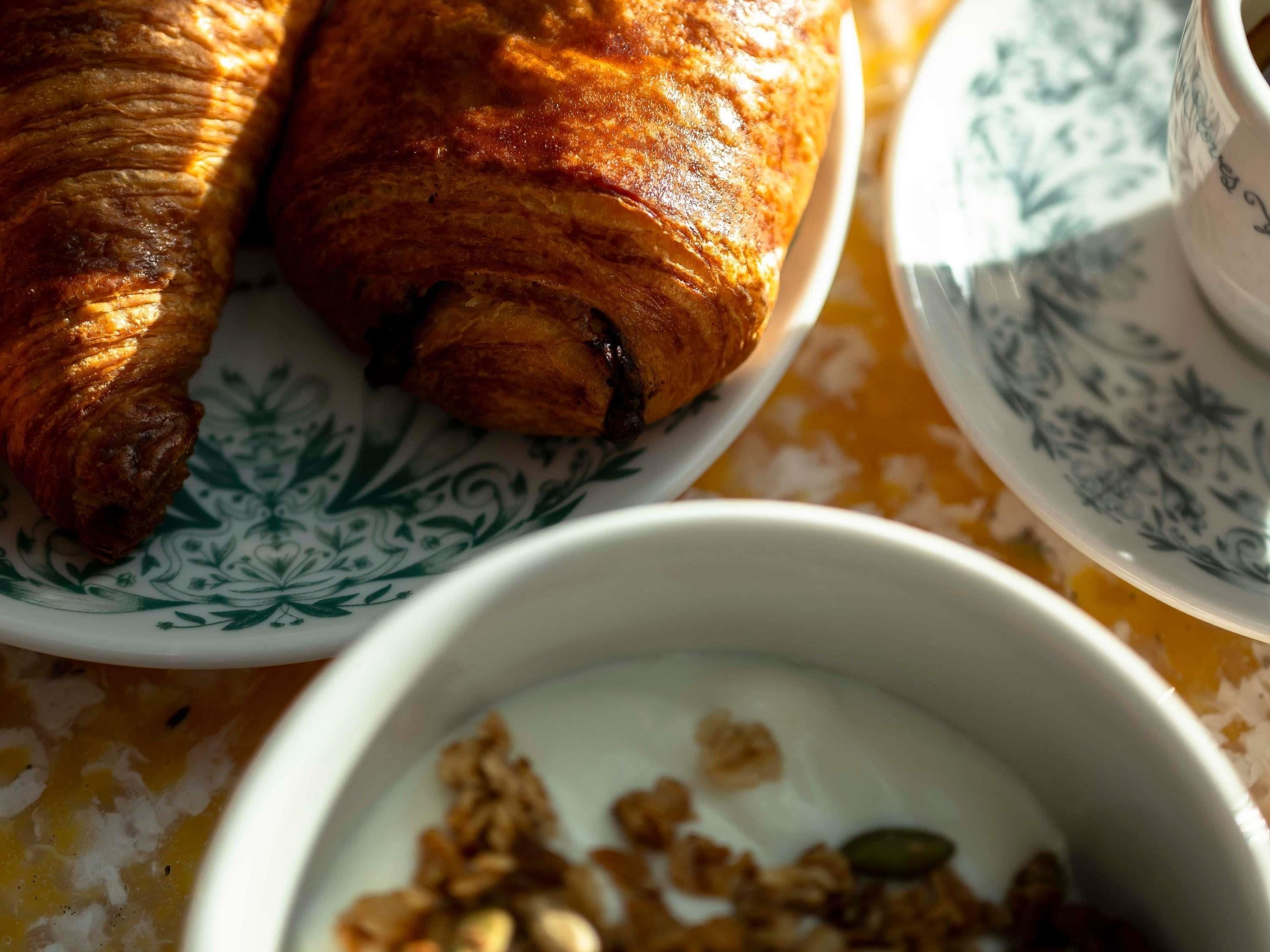Dos croissants sobre un plato estampado, un tazón de yogur con granola y una taza sobre una mesa.