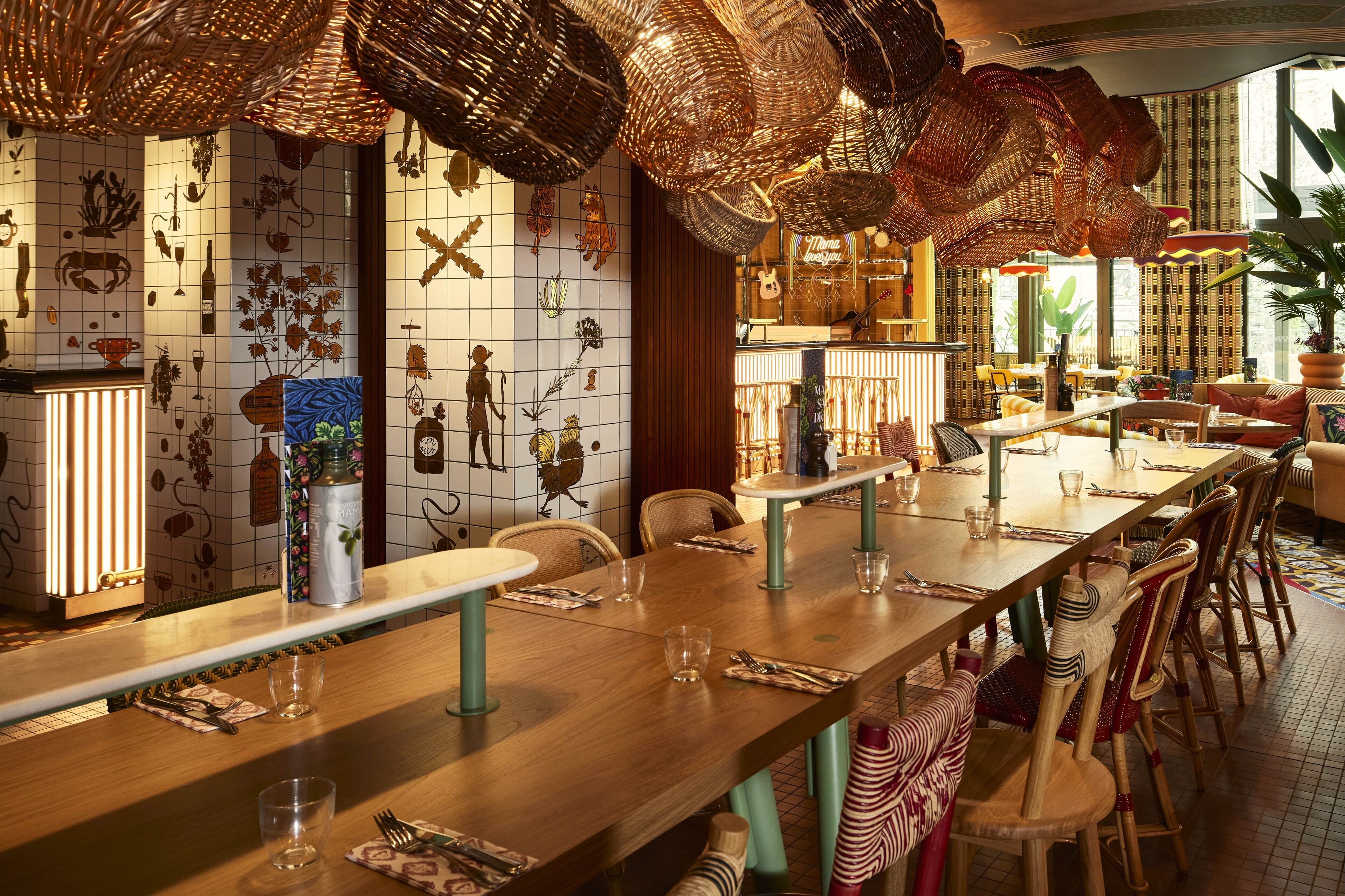 A restaurant with long wooden tables, wicker hanging lamps, and a tiled wall with various decorative elements.