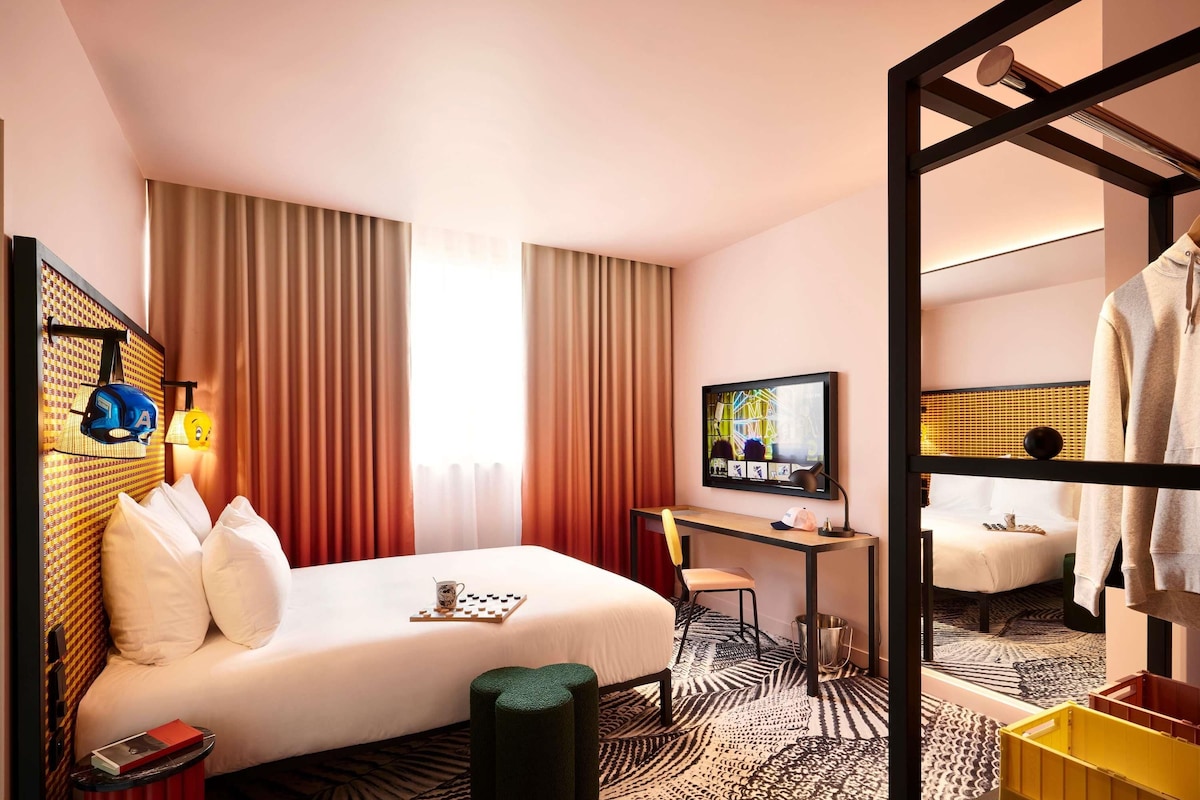 A hotel room with a bed, desk, TV, and a mirror reflecting the room.