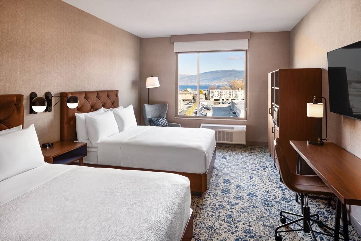 A hotel room with two beds, a desk, a chair, a TV, and a large window with a mountain view.