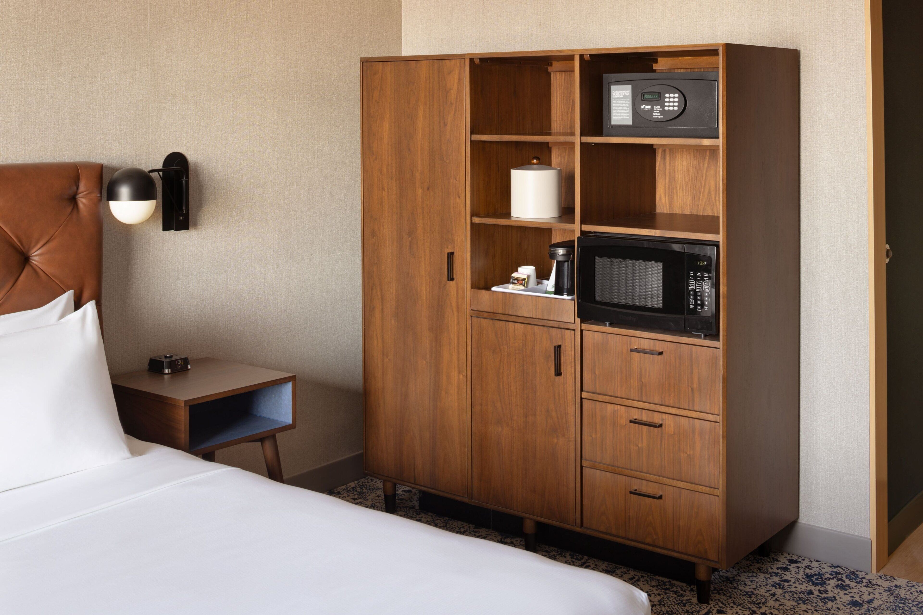 A hotel room with a bed, a wooden wardrobe, a bedside table with a lamp, and a microwave.