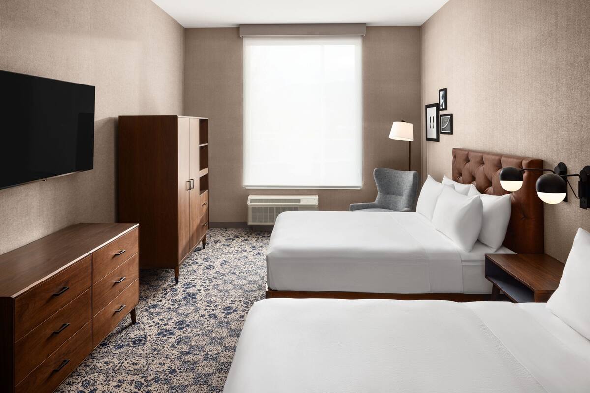 A hotel room with a large flat-screen TV, a wooden wardrobe, a bed with white bedding, a bedside table with lamps, and a window with blinds.