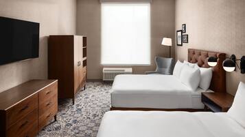 A hotel room with a large flat-screen TV, a wooden wardrobe, a bed with white bedding, a bedside table with lamps, and a window with blinds.