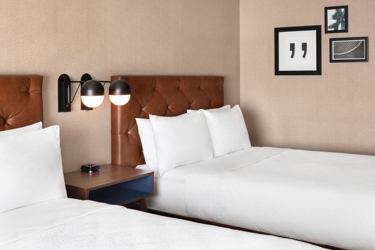 A hotel room with two beds, a nightstand, a lamp, and framed pictures on the wall.