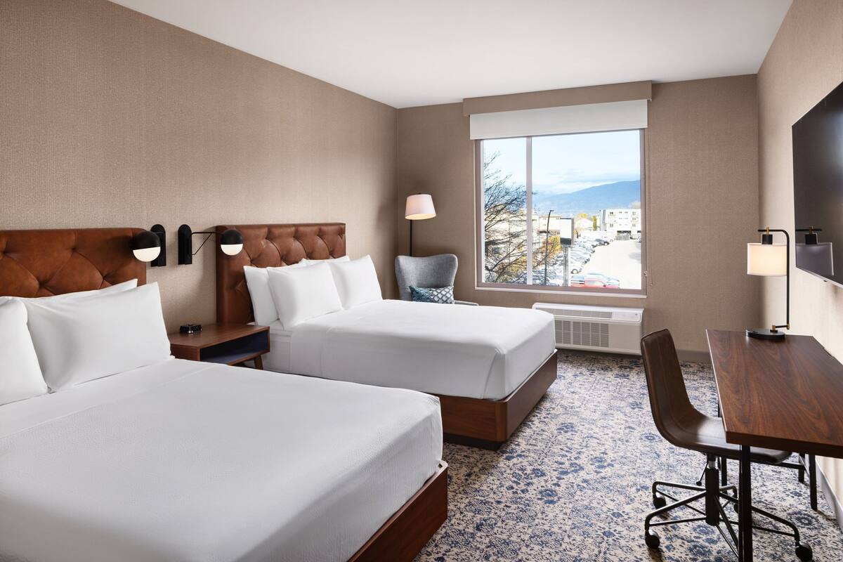 A hotel room with two beds, a desk, a chair, a large window with a view, and a flat-screen TV.