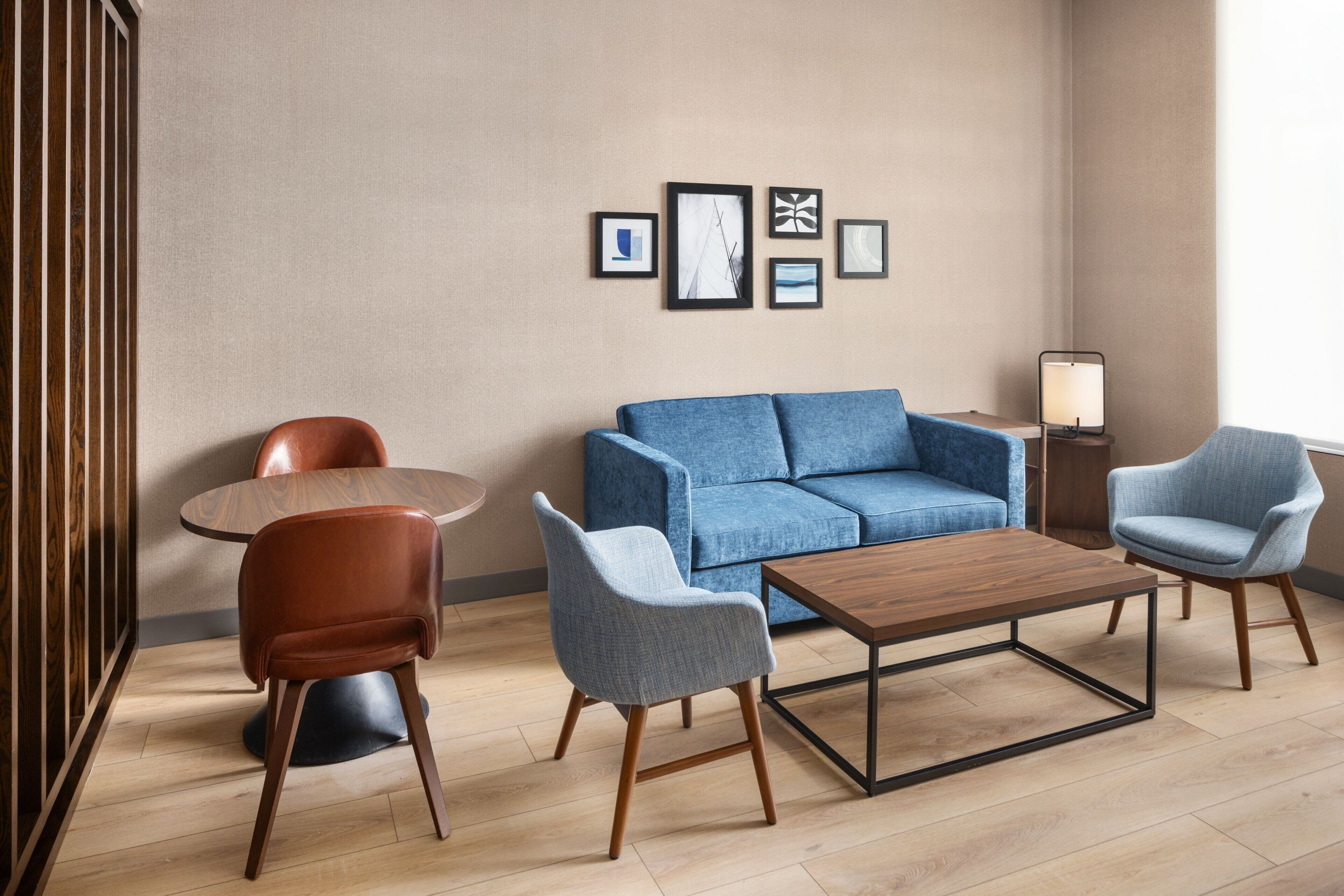 A modern living room with a blue sofa, two armchairs, a wooden coffee table, and a round table. There are framed pictures on the wall and a floor lamp.