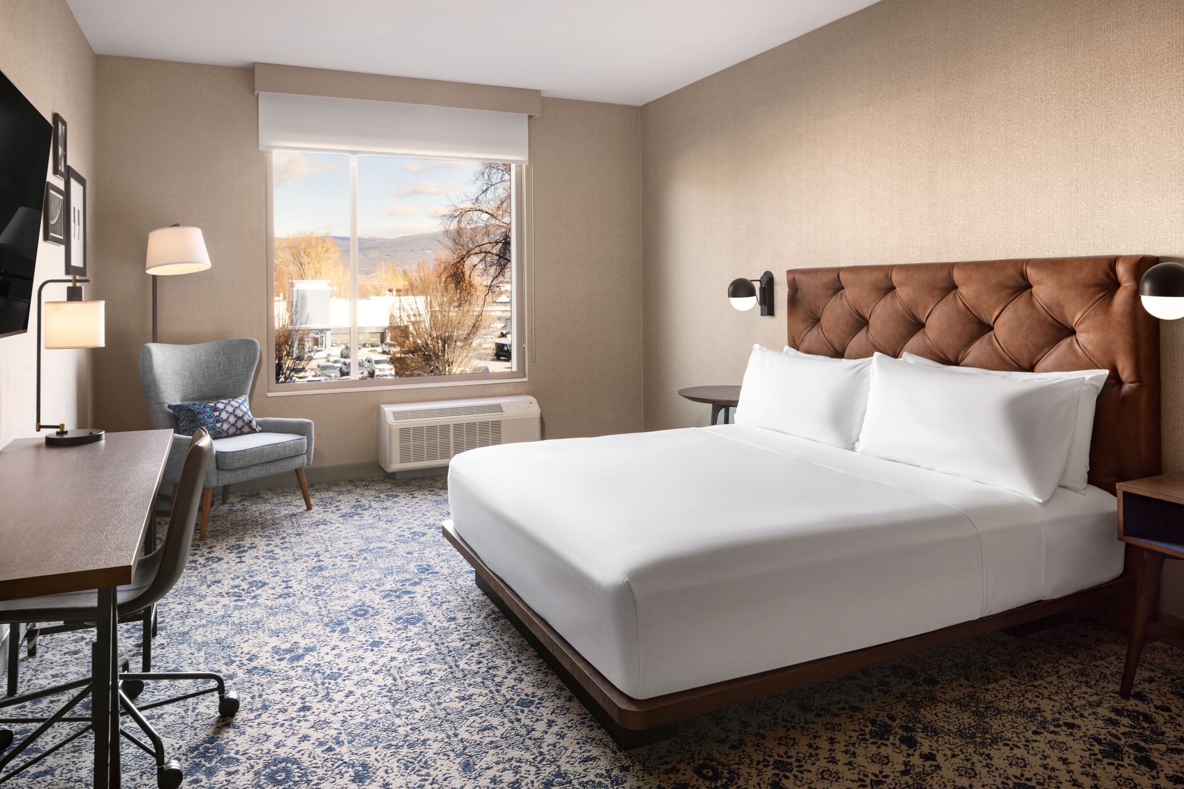 A hotel room with a bed, a desk, a chair, a window with a view, and a lamp.