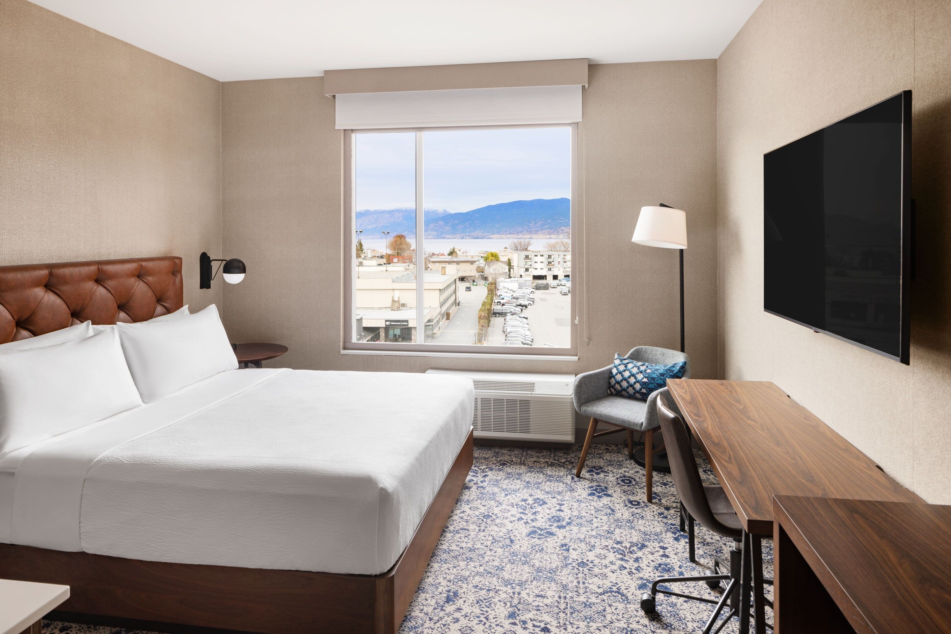 A hotel room with a large bed, a desk, a chair, a TV, and a window with a city view.