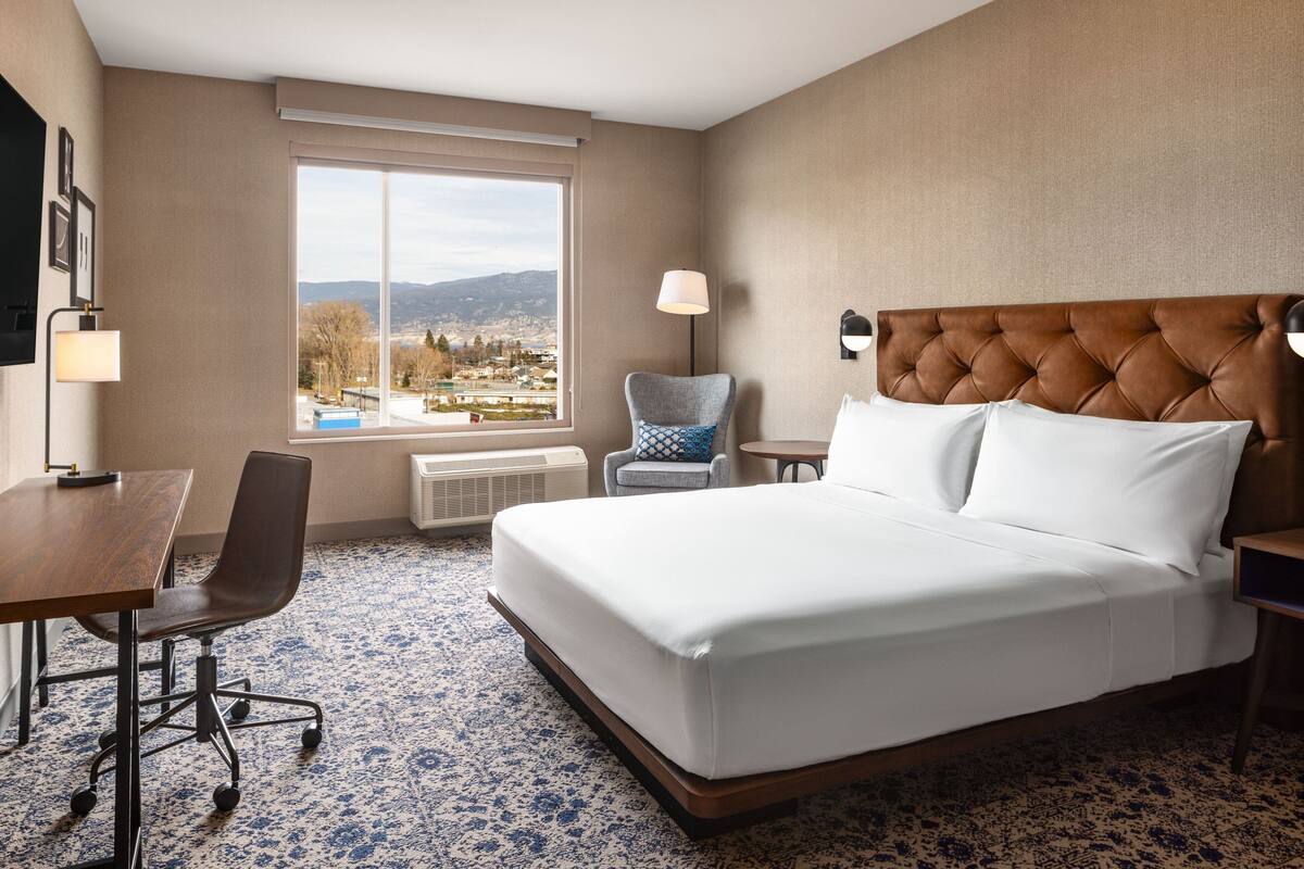 A hotel room with a large bed, a desk, a chair, and a view of a cityscape through a window.
