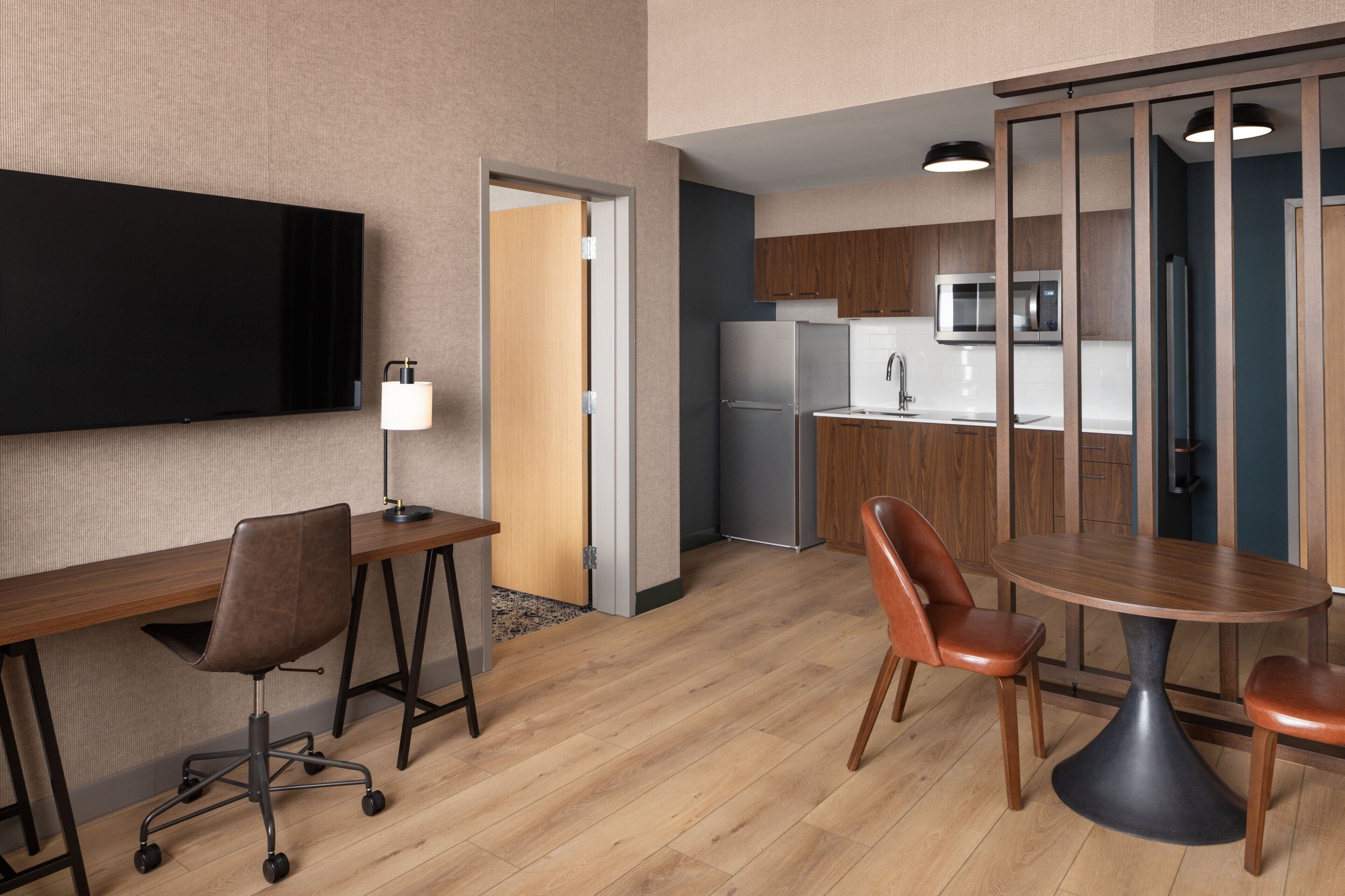 Habitación de hotel moderna con escritorio de madera, silla de oficina y mesa de comedor con sillas. Hay un televisor de pantalla plana en la pared, un microondas y un refrigerador en la zona de la cocina.