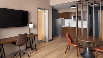 Habitación de hotel moderna con escritorio de madera, silla de oficina y mesa de comedor con sillas. Hay un televisor de pantalla plana en la pared, un microondas y un refrigerador en la zona de la cocina.