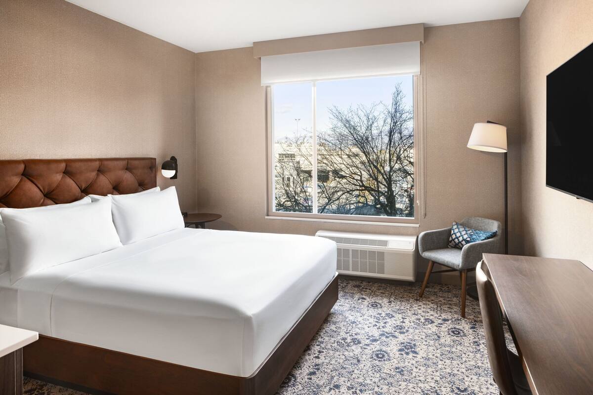 A hotel room with a large bed, a chair, a desk, a window with a view, and a flat-screen TV.