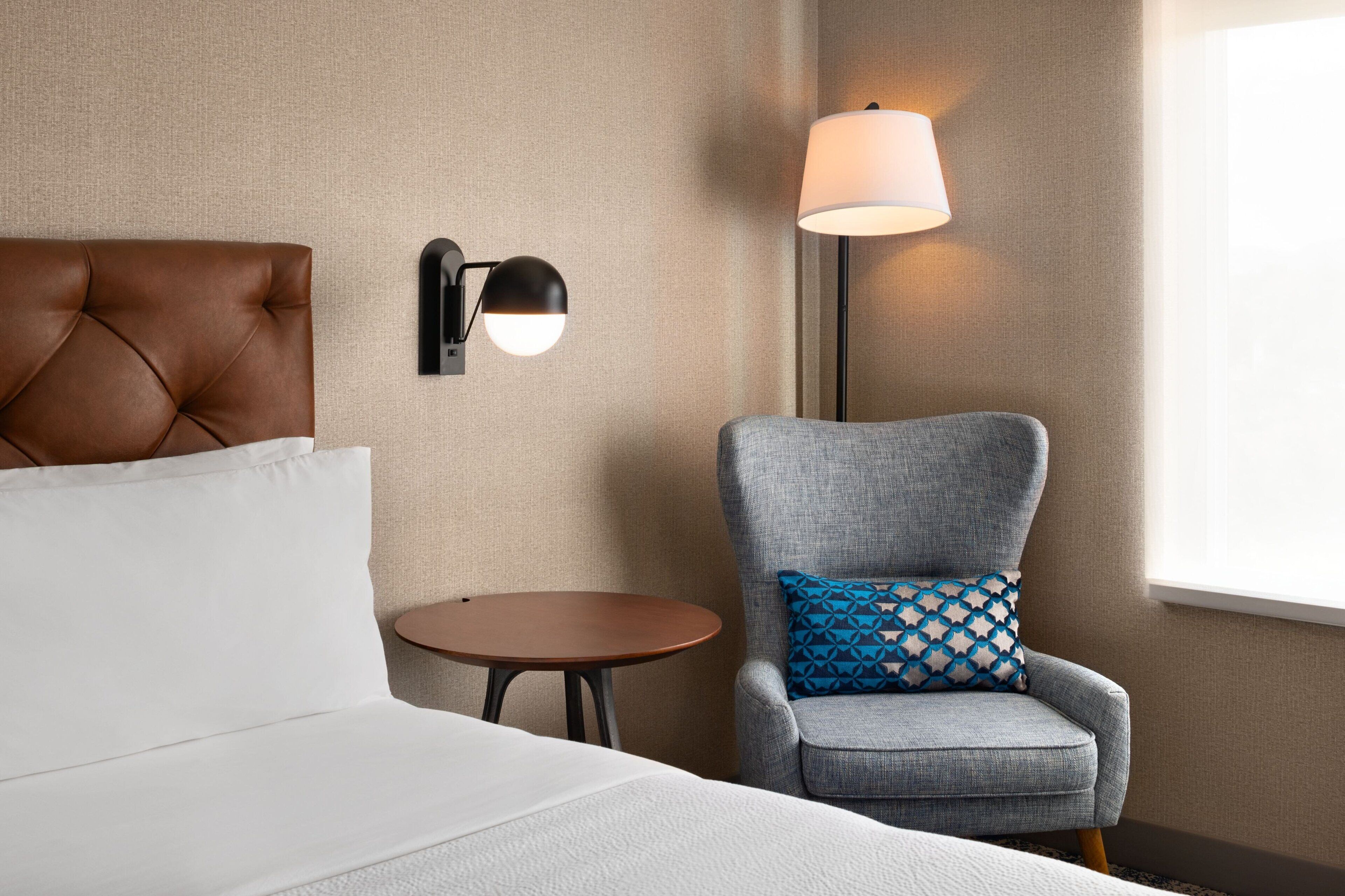 A hotel room with a tufted headboard bed, a grey armchair with a patterned pillow, a small round table, and two wall-mounted lamps.