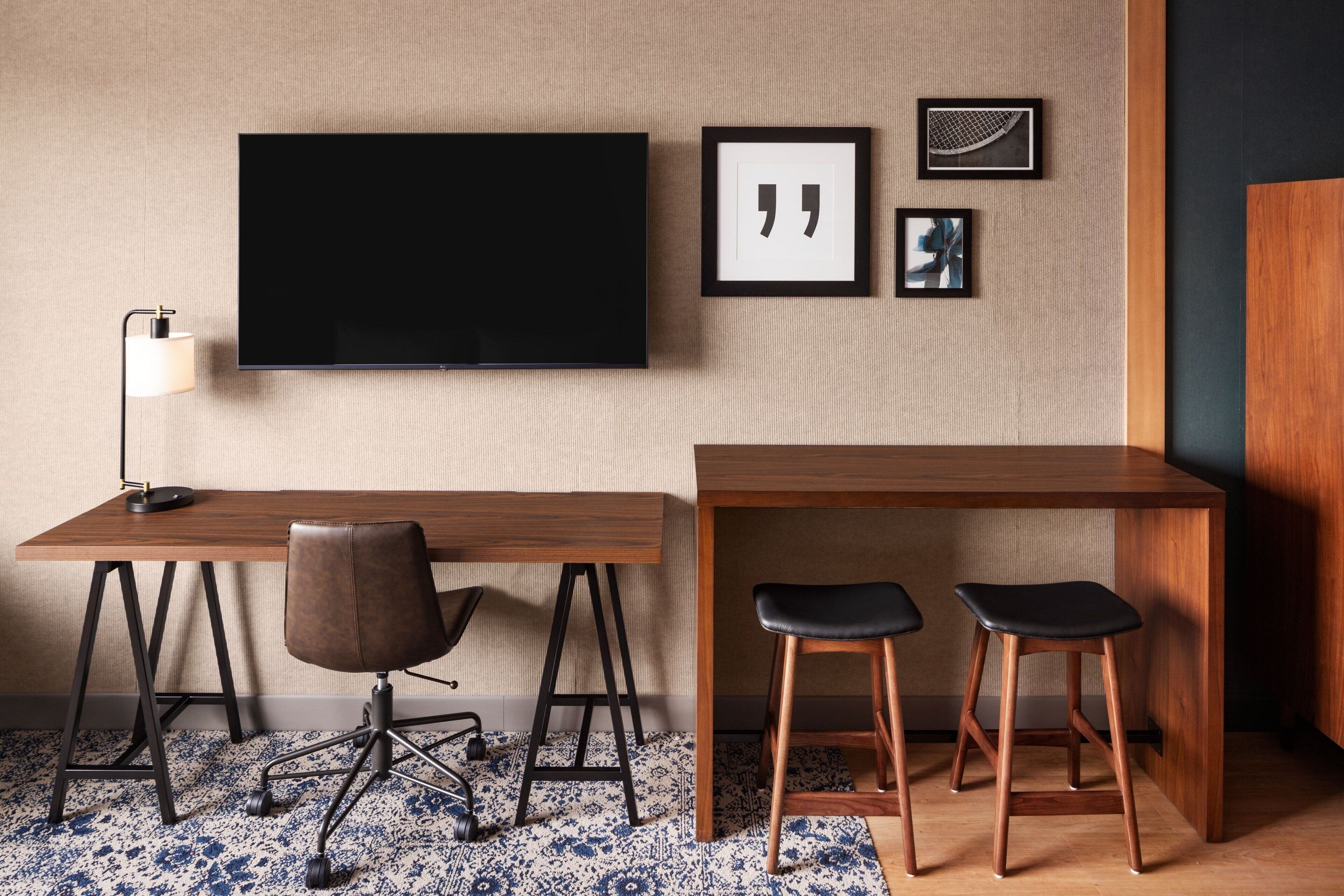 A modern hotel room with a large flat-screen TV mounted on the wall, a wooden desk with two black bar stools, a brown leather chair, and a patterned rug.