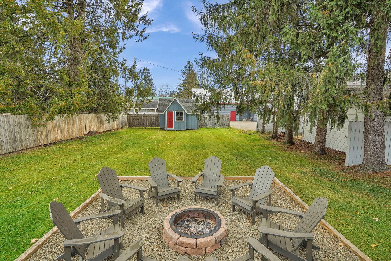 A backyard with a fire pit and Adirondack chairs, surrounded by a wooden fence and trees.