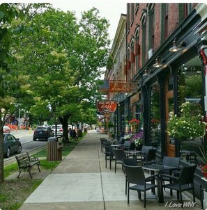 Un café avec terrasse donnant sur le trottoir, un bâtiment en briques avec une devanture de magasin, et une rue bordée de voitures garées.
