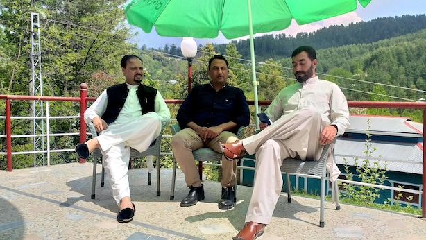 Three men seated on outdoor chairs under a green umbrella, with a mountainous backdrop. - Banjosa Inn Guest House (Chhota Gala)