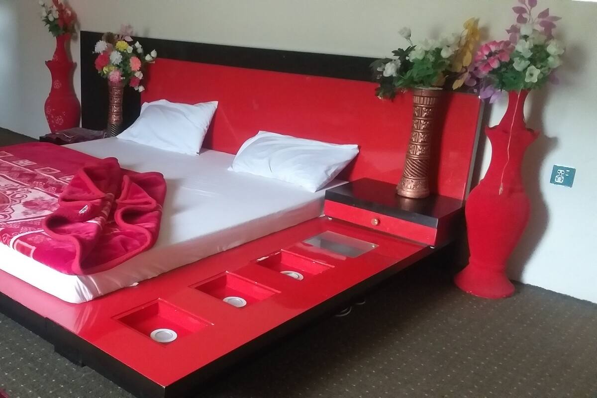 Uma cama com colcha vermelha e branca, duas almofadas brancas e cabeceira vermelha. Há duas mesas de cabeceira vermelhas com vasos de flores.