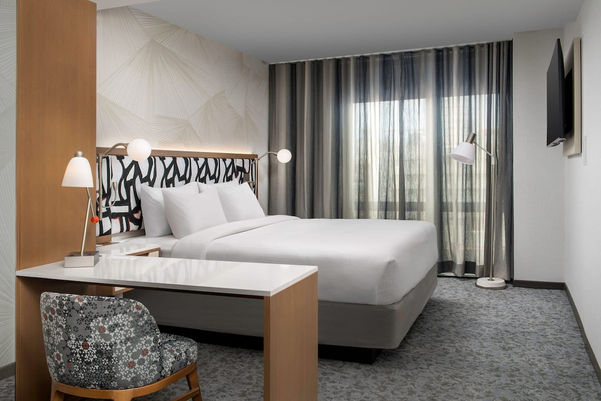 A hotel room with a bed, a desk with a chair, a TV mounted on the wall, and a window with curtains.