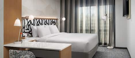 Quarto de hotel com cama, escrivaninha com cadeira, televisão na parede e janela com cortinas.