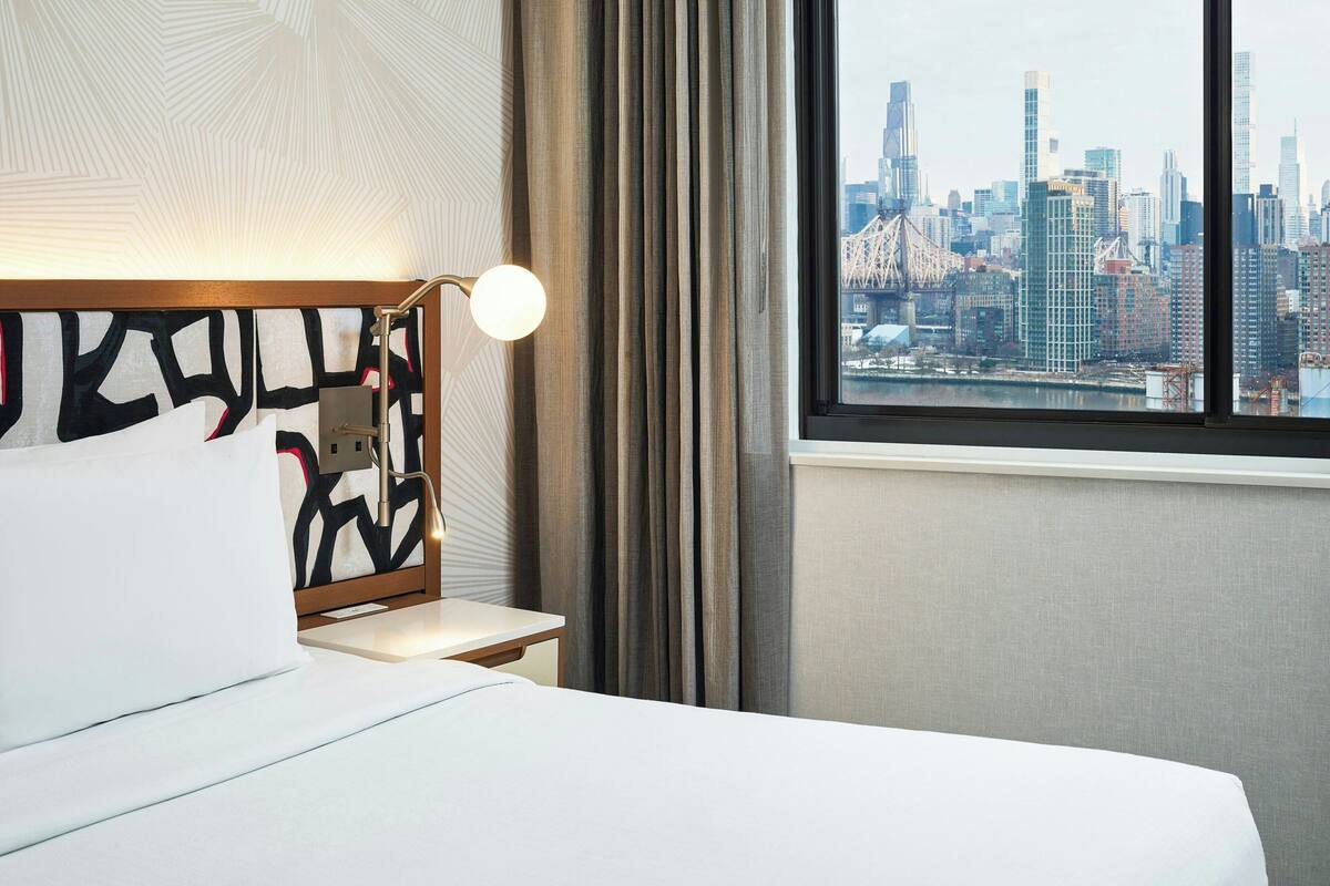 A hotel room with a bed, a bedside table with a lamp, a view of a city skyline through a window.