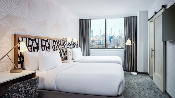 A hotel room with a bed, a desk, a chair, a lamp, and a view of the city skyline. - SpringHill Suites by Marriott New York Queens (Long Island City)