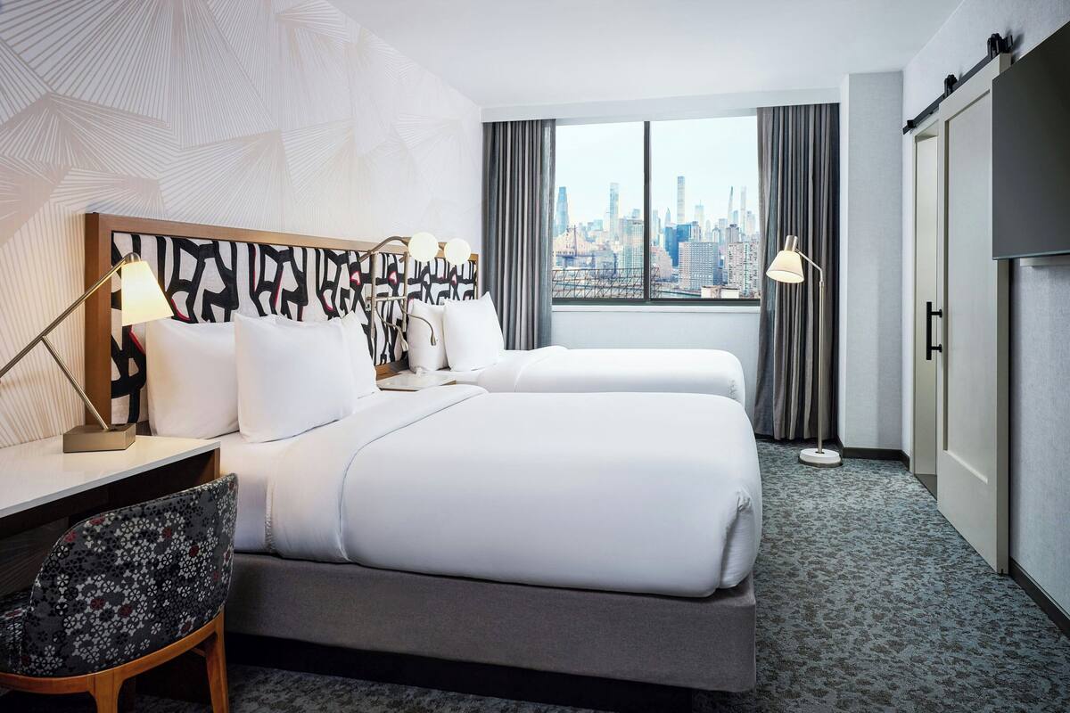 A hotel room with a bed, a desk, a chair, a lamp, and a view of the city skyline.