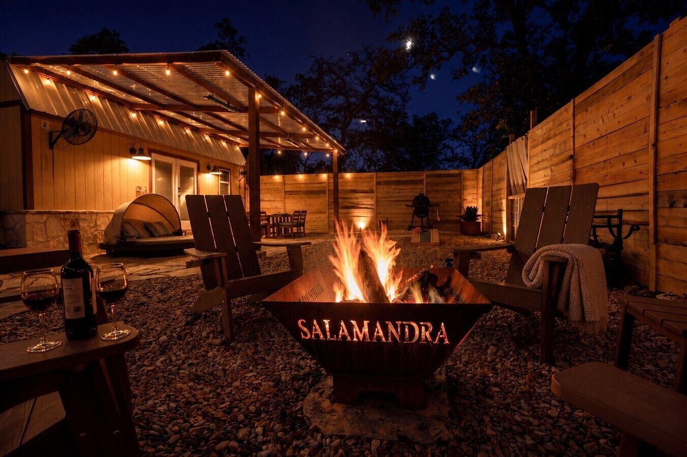 Ein Außenbereich mit Terrassenmöbeln, einer Feuerstelle mit der Aufschrift 'SALAMANDRA' und einem Holzgebäude.
