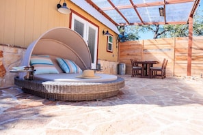 A cozy outdoor seating area with a curved wicker sofa, a dining table with chairs, and a pergola overhead. - A+ Privacy | Romantic Lux | Sauna | Hot/Cold Pool (Wimberley)