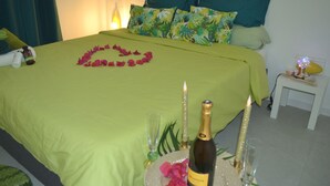 Een net opgemaakt bed met groen beddengoed, een bloemstuk in de vorm van een hart, een fles champagne en twee brandende kaarsen op een dienblad.