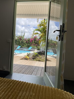 On aperçoit, à travers une porte vitrée, un jardin avec une piscine, une terrasse en bois et différentes plantes.