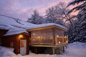 Uma cabana de madeira com varanda envidraçada, rodeada por árvores cobertas de neve.
