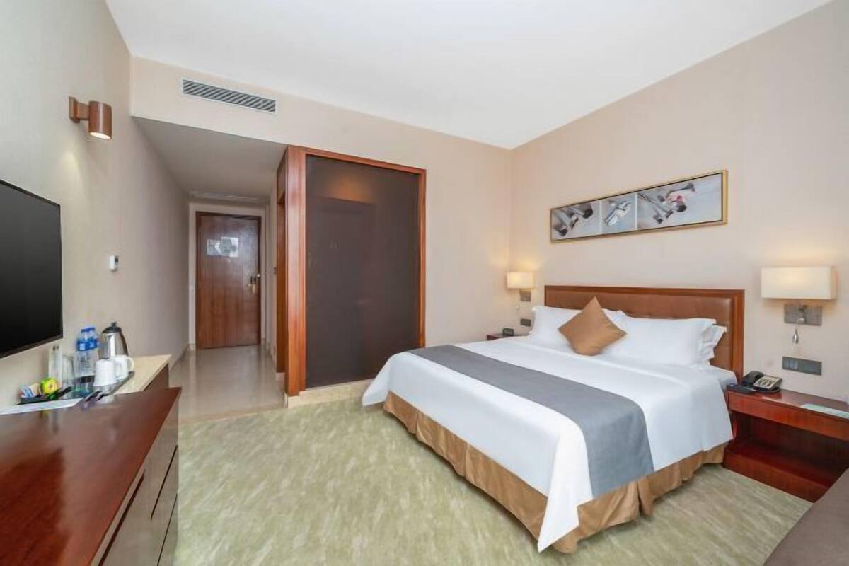 A hotel room with a bed, bedside lamps, a TV, a desk with a coffee maker, and a door leading to another room.