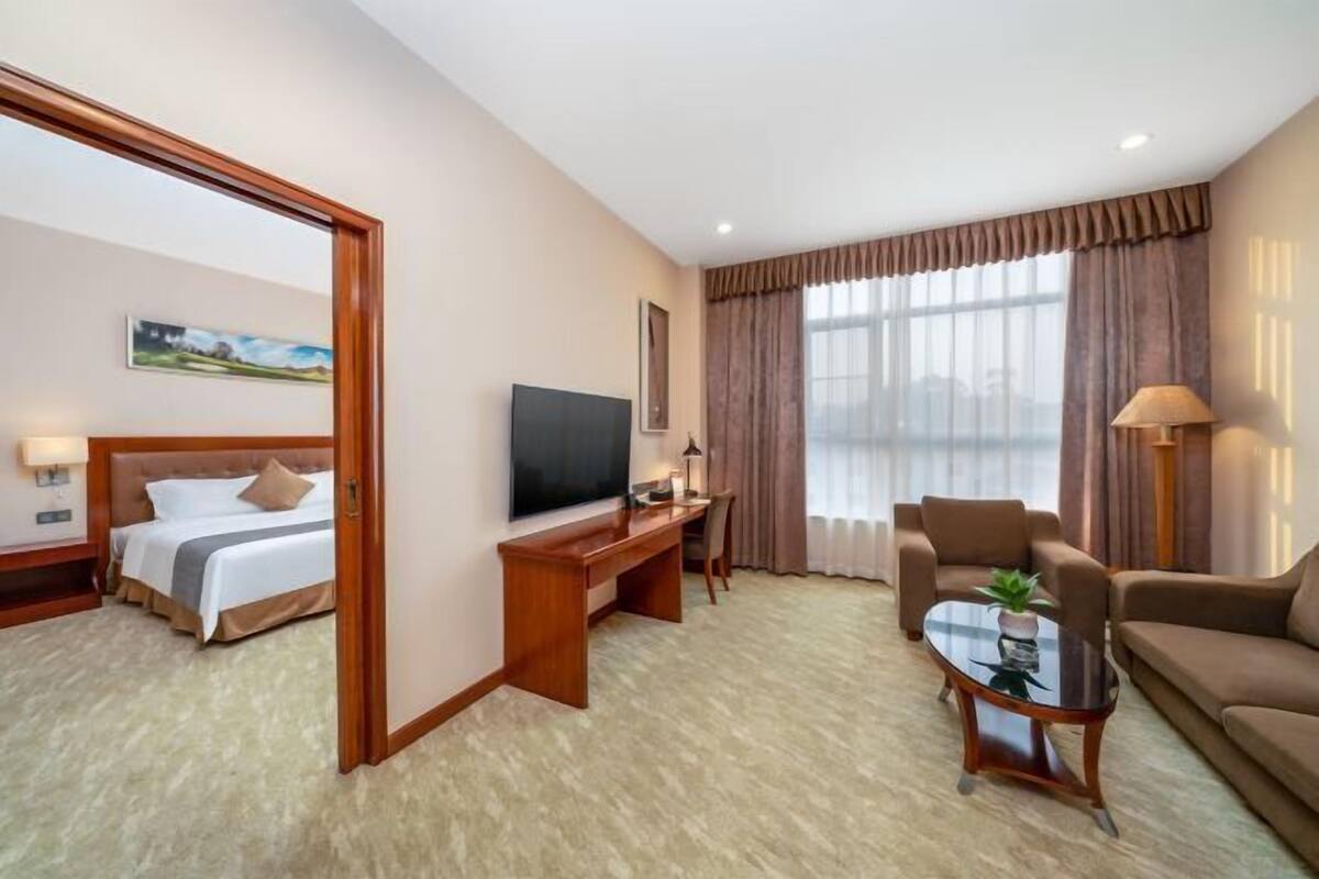 A hotel room with a bed, a TV, a desk, a chair, a sofa, and a large window with curtains.