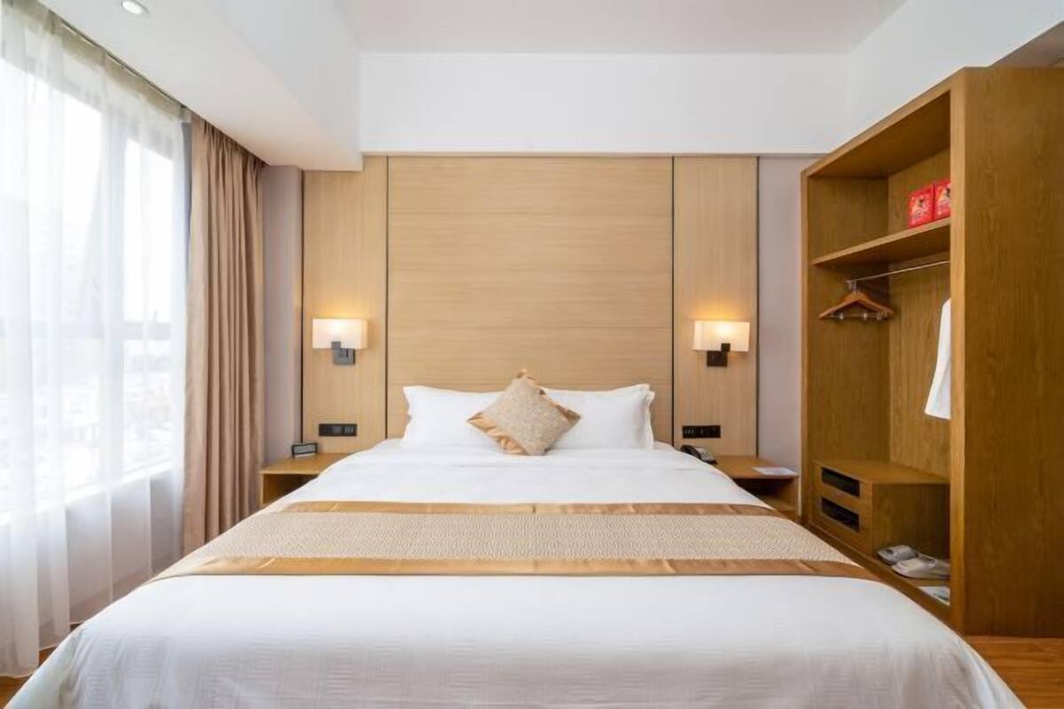A hotel room with a large bed, wooden headboard, bedside lamps, a wardrobe, and a view of the city.