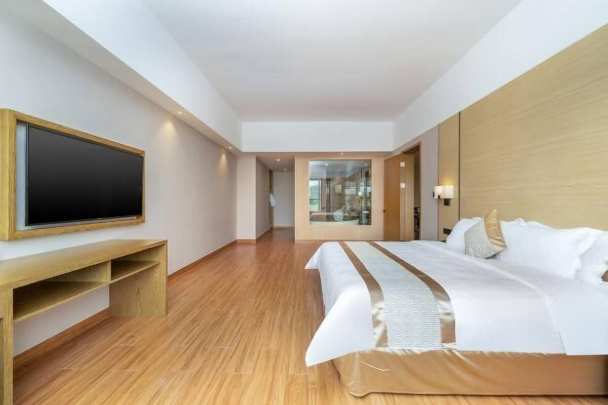 A modern hotel room with a large bed, a flat-screen TV, a wooden bench, and a glass door leading to a balcony.