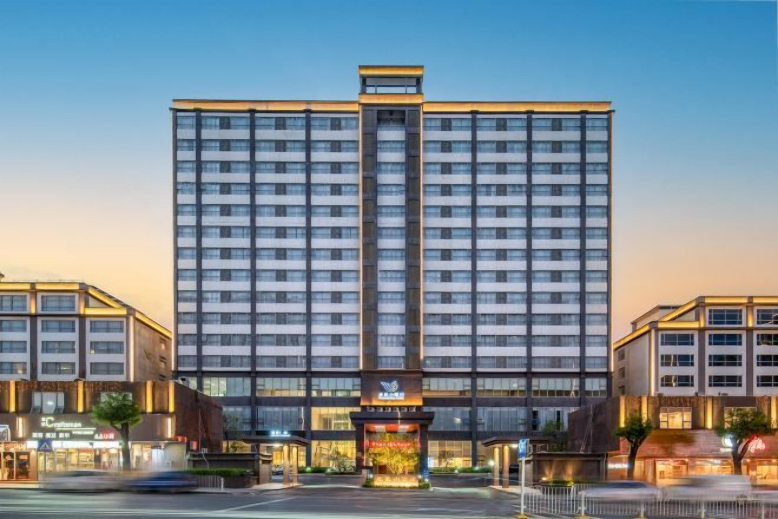 Un hotel de varios pisos con una entrada central, rodeado de edificios más pequeños, y un cielo despejado.