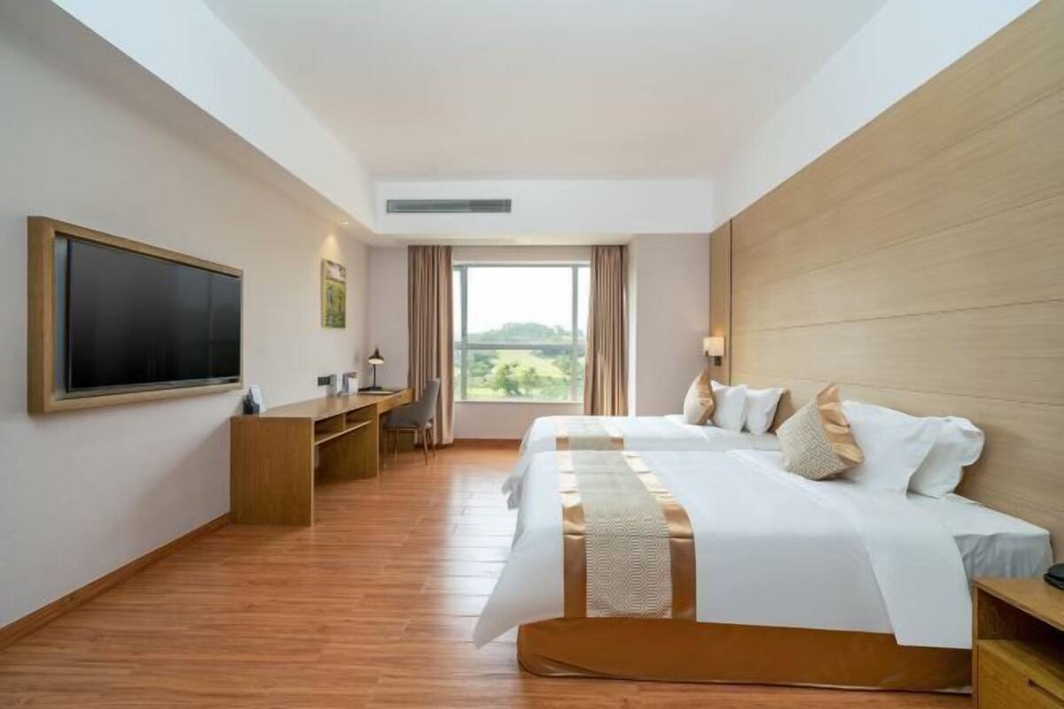 A hotel room with a large bed, a desk, a flat-screen TV, and a window with a view of greenery.