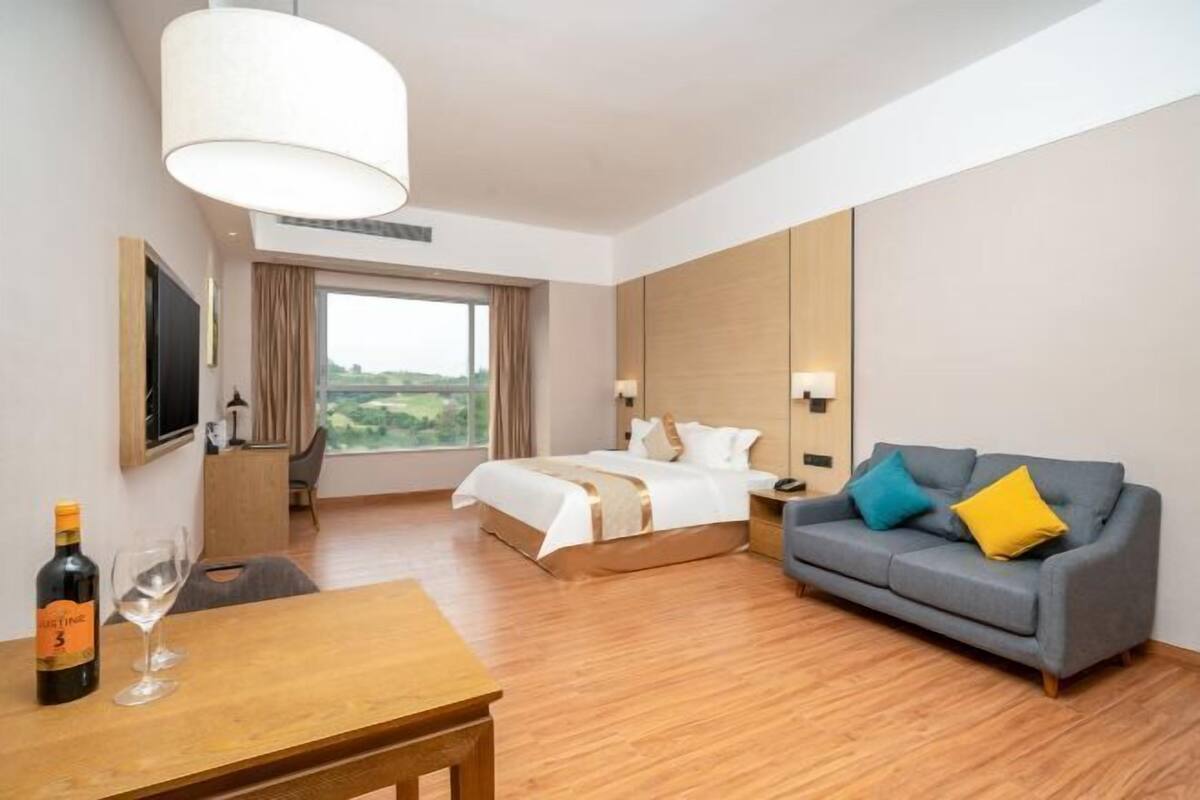 A hotel room with a bed, a sofa, a table with a bottle and glass, and a large window.