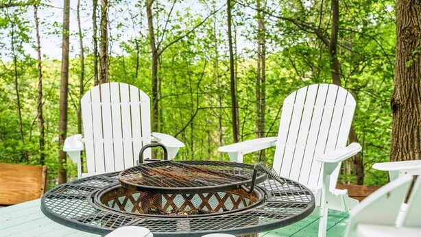 Een ronde tafel met een grill in het midden, omringd door witte Adirondack-stoelen, geplaatst op een houten terras met een bos op de achtergrond.