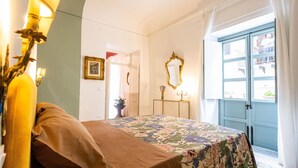 A bedroom with a floral bedspread, a bedside table, a mirror, and a view of the outside through a door.