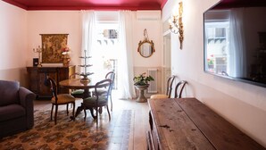 A room with a dining table, chairs, a sofa, a chandelier, a mirror, and a window with curtains.