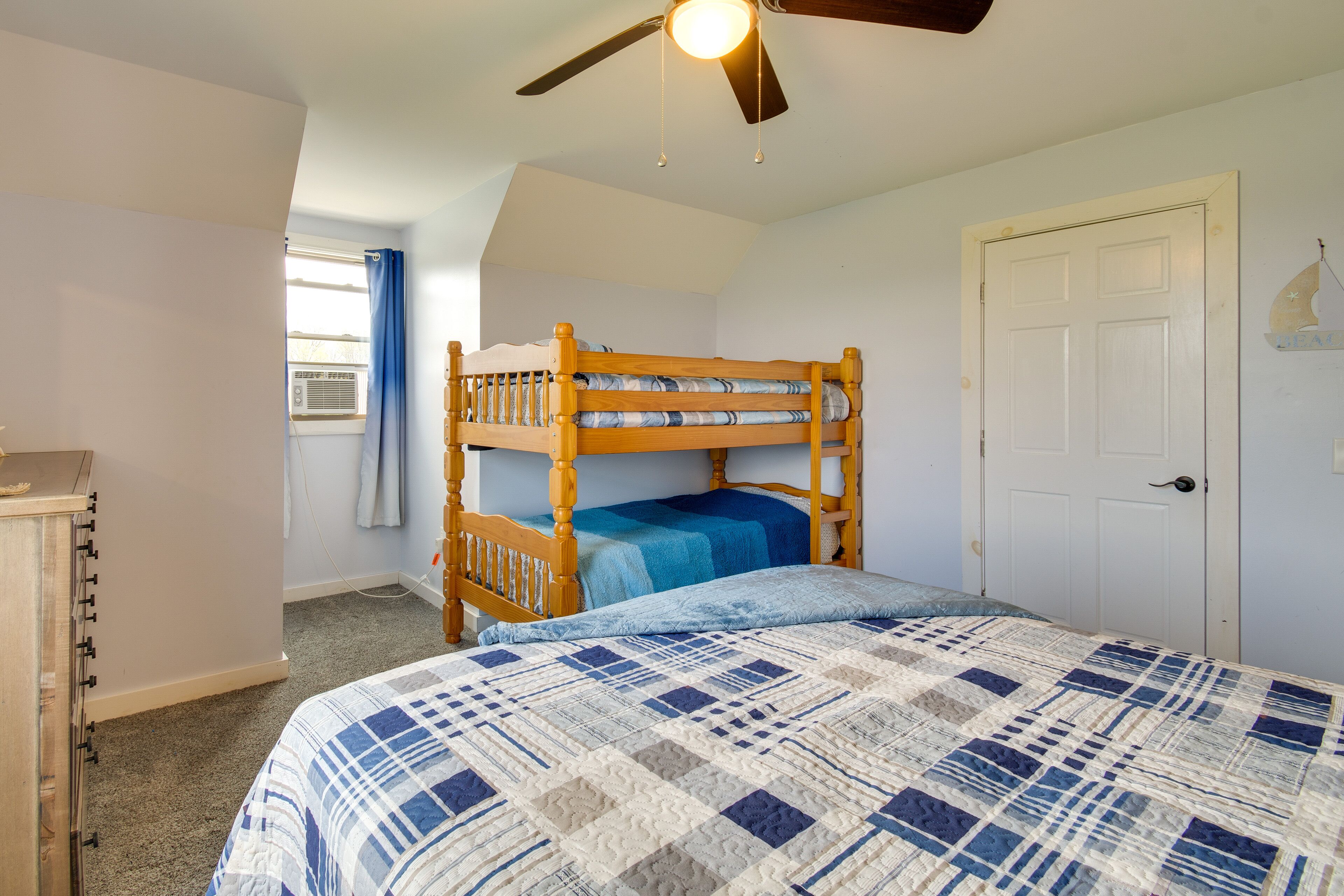 Ein Etagenbett mit einer blau-weißen Steppdecke, ein Deckenventilator, eine weiße Tür und ein Fenster mit blauen Vorhängen.
