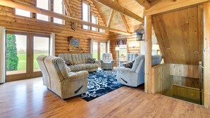 L’intérieur d’une cabane en rondins, avec un plafond en bois, de grandes fenêtres, un salon doté de canapés et de fauteuils, et une vue sur l’extérieur.