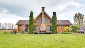 Une cabane en rondins avec une cheminée en pierre, de grandes fenêtres et une véranda dotée de sièges d’extérieur.