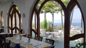 A dining area with tables set for a meal, large arched windows, and a view of the ocean. - Hotel Parco Conte (Casamicciola Terme)