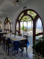 Un comedor con mesas dispuestas para una comida, amplias ventanas en arco y vista al océano.