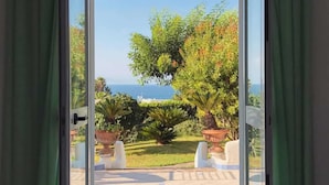 A view through a glass door of a garden with a patio area, a large urn, and a view of the sea. - Hotel Parco Conte (Casamicciola Terme)